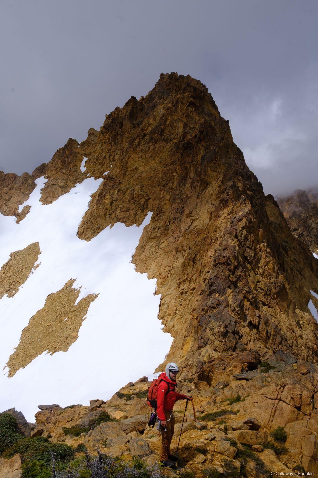 A rust-orange rock pyramid above a snowfield, a climber in red at its base