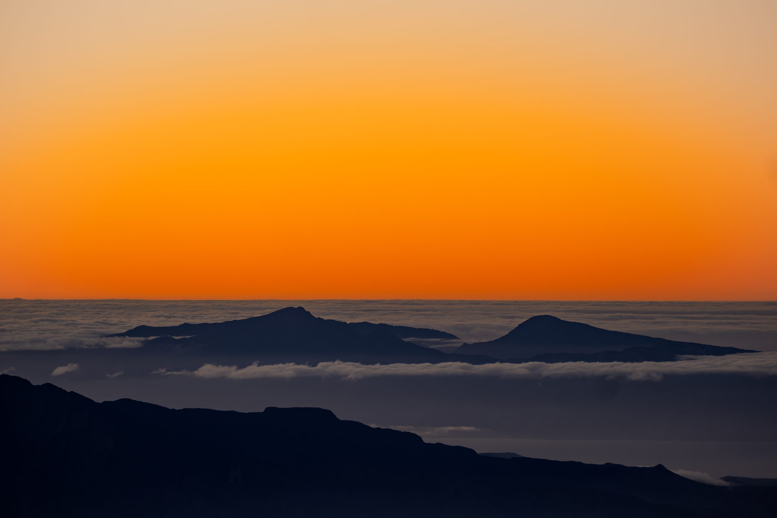Tangerine dawn sky over a sea of clouds, two distant volcanic summits breaking through.